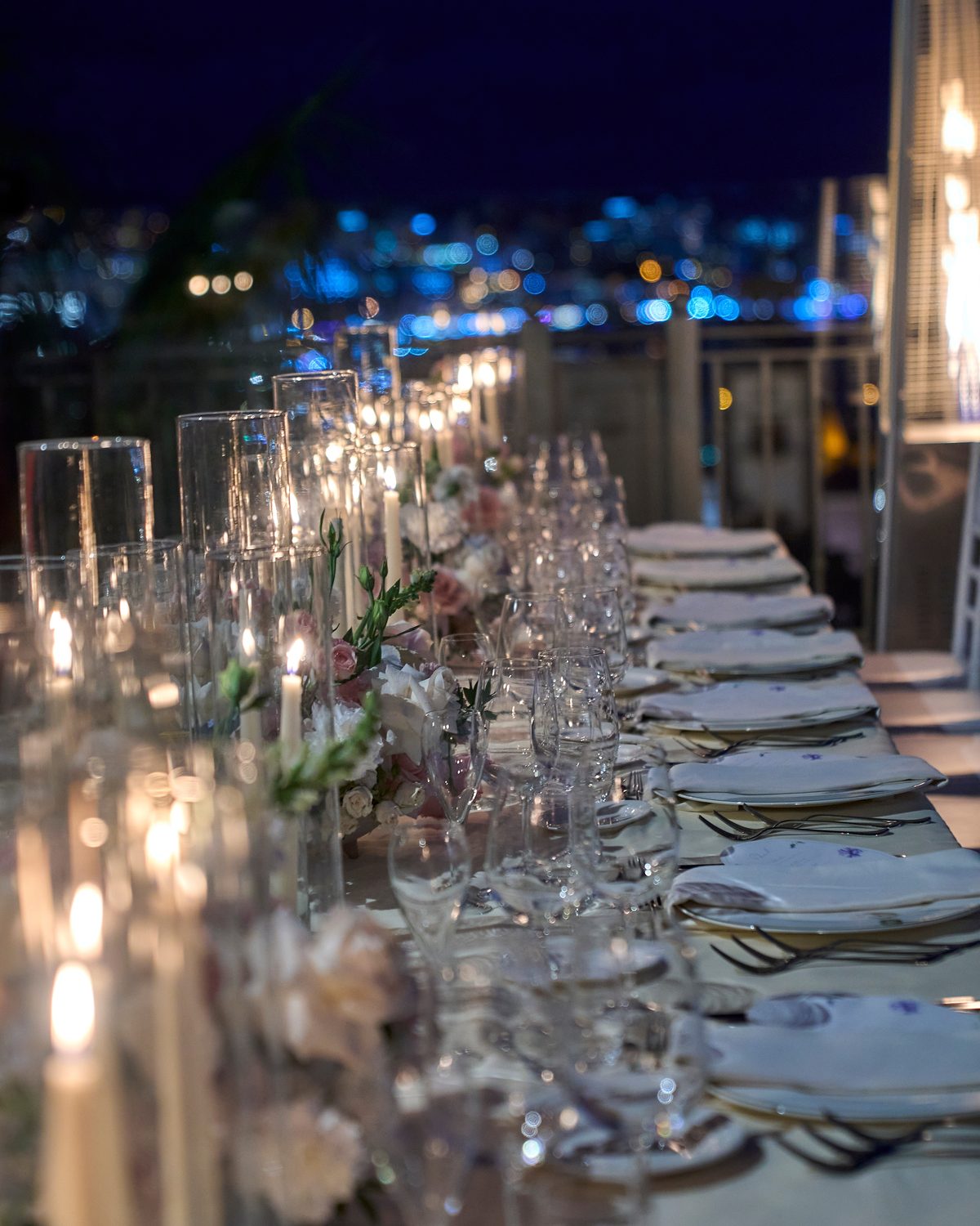 A long candlelit banquet table set for a private event at dusk, city lights beyond
